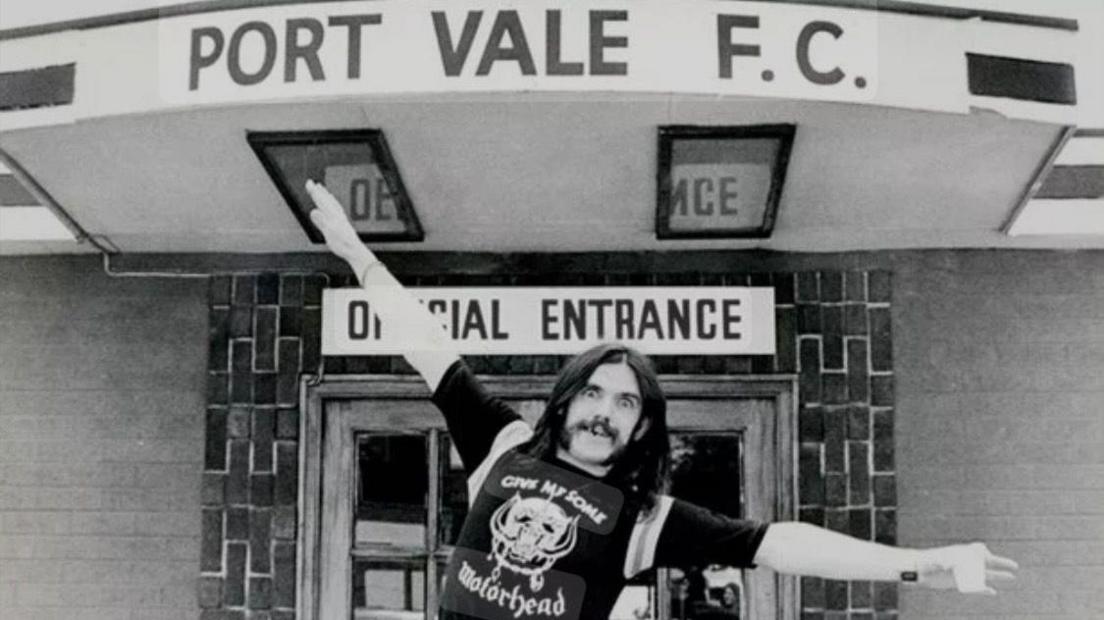 A black and white photograph of Motörhead frontman Lemmy, a man with long dark hair and a dark moustache, holding his arms out like a plane underneath a sign reading "PORT VALE F.C.". Another sign under that reads "OFFICIAL ENTRANCE".