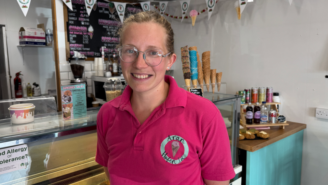 A woman with tied-back blonde hair and glasses, wearing a pink polo shirt and smiling as she looks at the camera. Behind her is an ice cream parlour front cabinet and worktops with pastries, drinks and a coffee machine on top.
