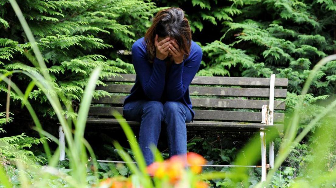 A woman on a bench surrounded by trees and plants holds her head in her hands