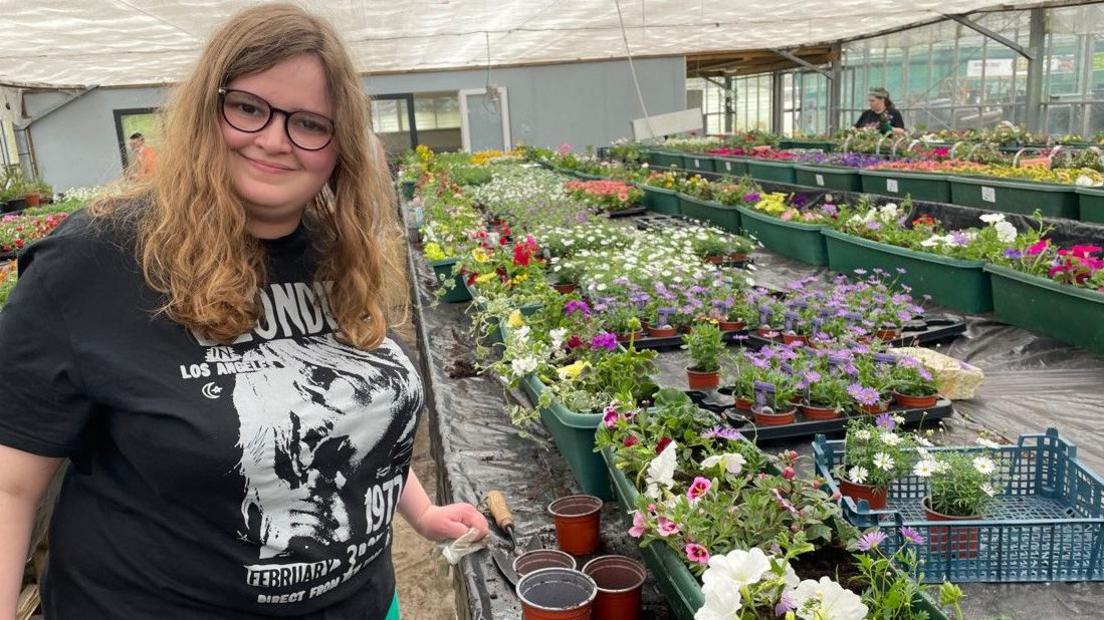 A smiling woman posing for the camera in a large glasshouse next to a stand full of different potted flowering plants. She has long brown hair and dark-rimmed glasses and is wearing a black Blondie band t-shirt.