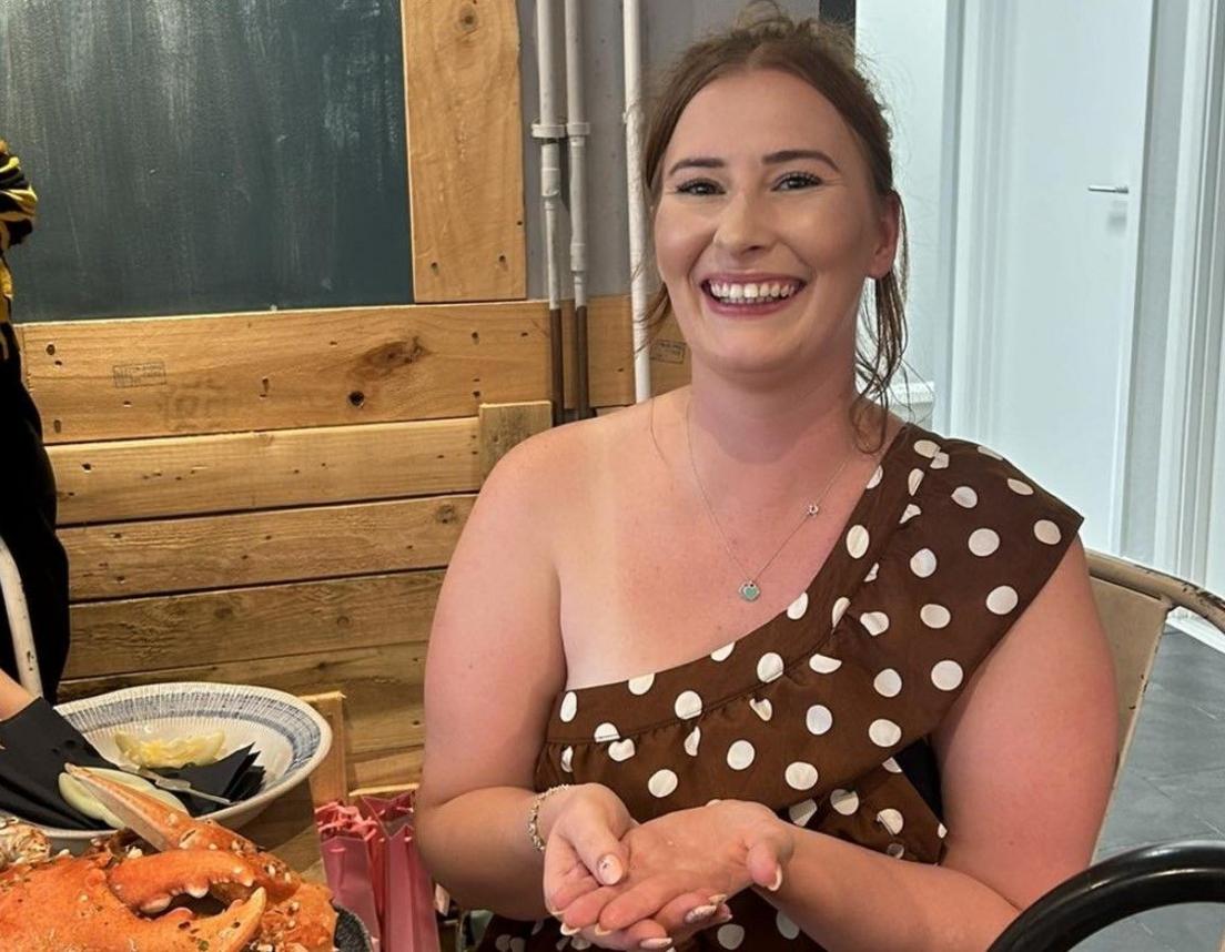 A woman in a brown dress with white polka dots sits smiling at a table with her palm held open. On the table to her left a crab's claw is visible