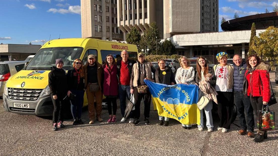 A large group of people, with some holding up a flag, standing by an ambulance, in Ukraine. They are all looking at the camera and smiling. A building is behind them. 