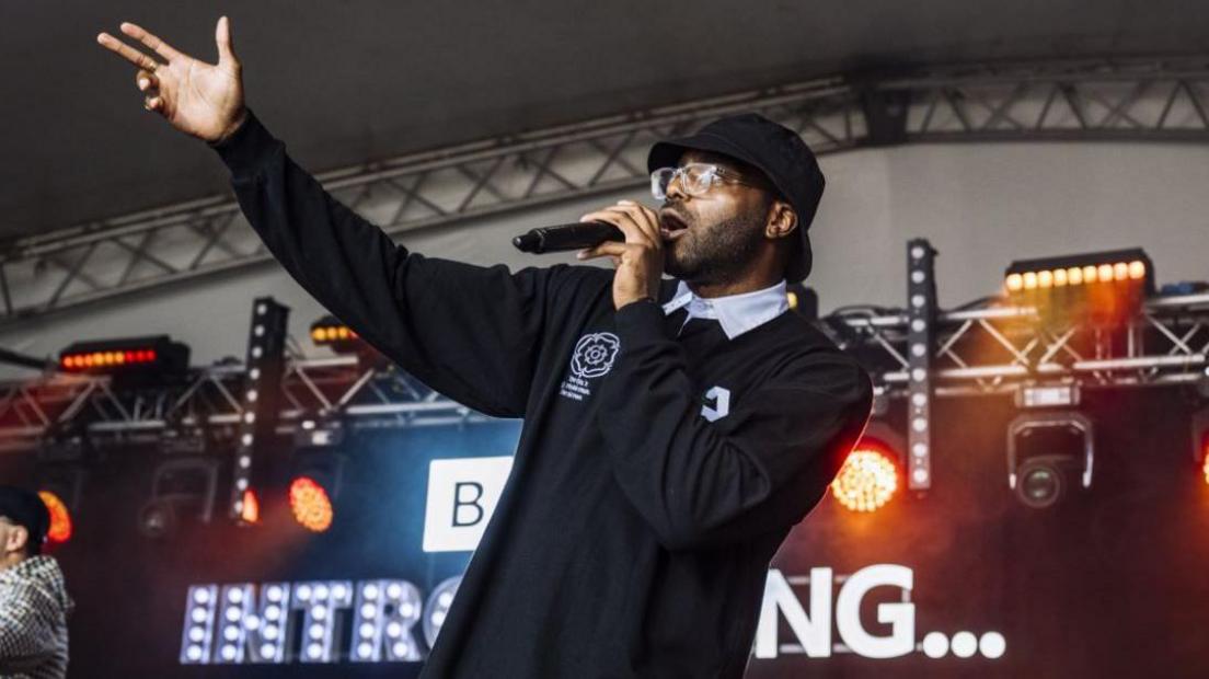 A man - Chiedu Oraka - on stage wearing a black long-sleeve polo shirt with a white collar and two white logos on either side on the chest. He is wearing a black bucket hat and clear-framed glasses. He is holding a black microphone to his mouth and is holding his right arm straight in the air. Red and orange lights can be seen in the background as well as a set of lights reading 'BBC INTRODUCING' in capital letters.