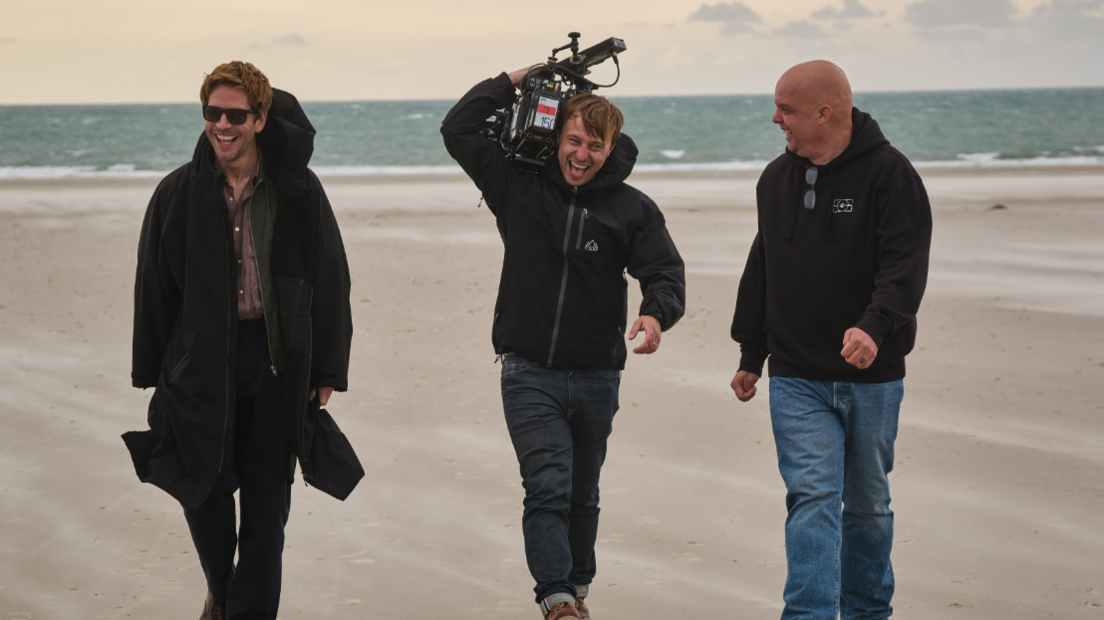 Damien Molony on the left wearing a long coat walking along a beach in St Ouen's Bayin. A man in the middle carrying film equipment and a third man on the right. 