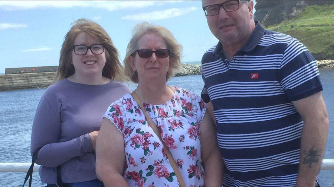 Emma, Audrey and Eddie standing huddled together smiling with the sea behind them as well as a green hill. The couple are in t-shirts and Emma is wearing a purple jumper. 