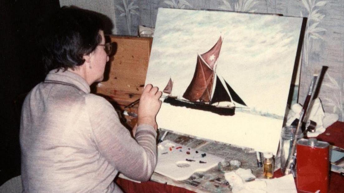 A woman painting a sail boat onto a canvas. She has her back turned to the camera and is wearing a beige jumper.