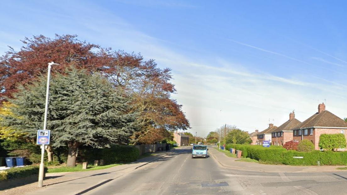 A quiet-looking road, bordered by 1950s houses and large trees. There is a junction on the right.