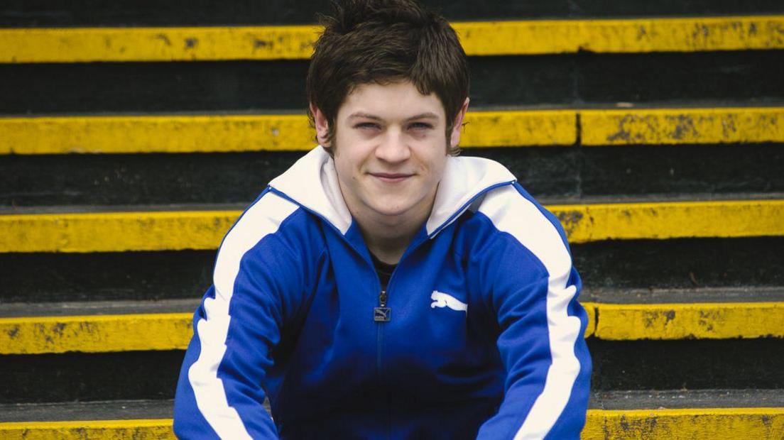 A young Iwan Rheon wearing a tracksuit smiles at the camera