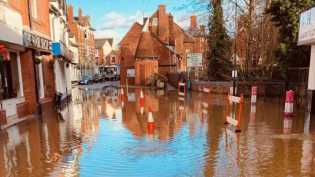 A flooded street in a town centre, with orange and white traffic cones and barriers placed in the waters. Redbrick buildings line the streets.
