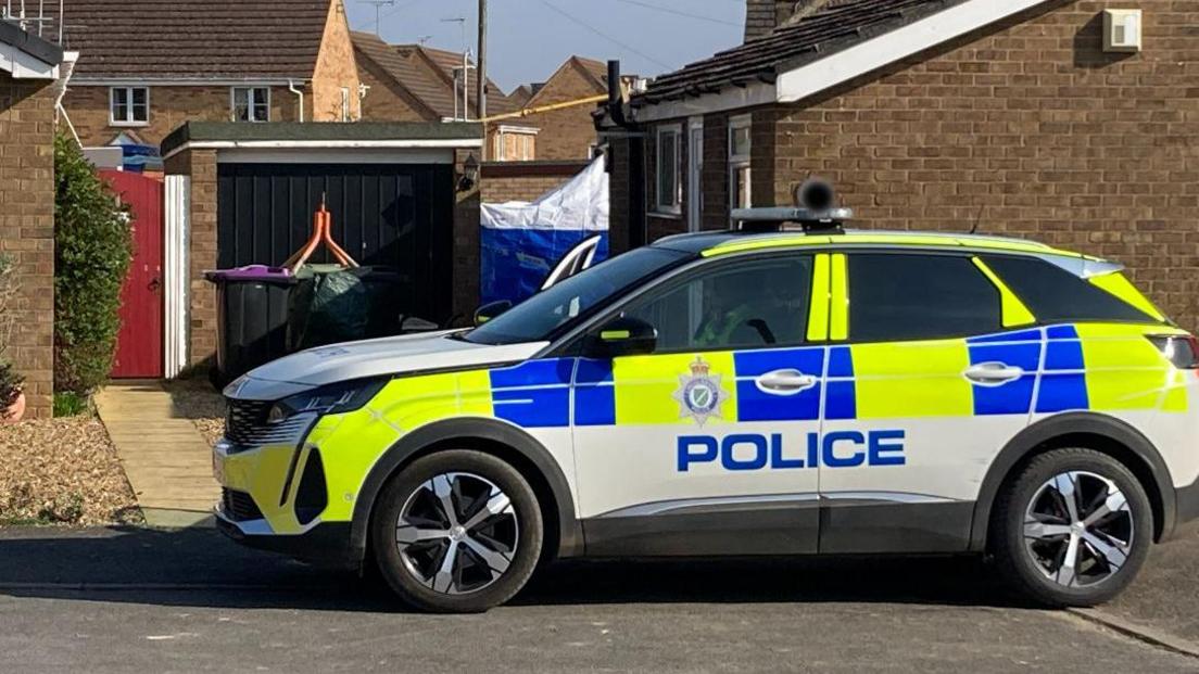 A police car is parked outside a bungalow.  You can see a police tent in the garden looking down a path at the side of the house.