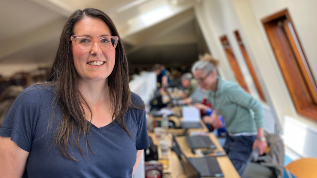 A woman with long dark hair, a dark blue t-shirt and pink-rimmed glasses smiles at the camera. Behind her are people using laptops