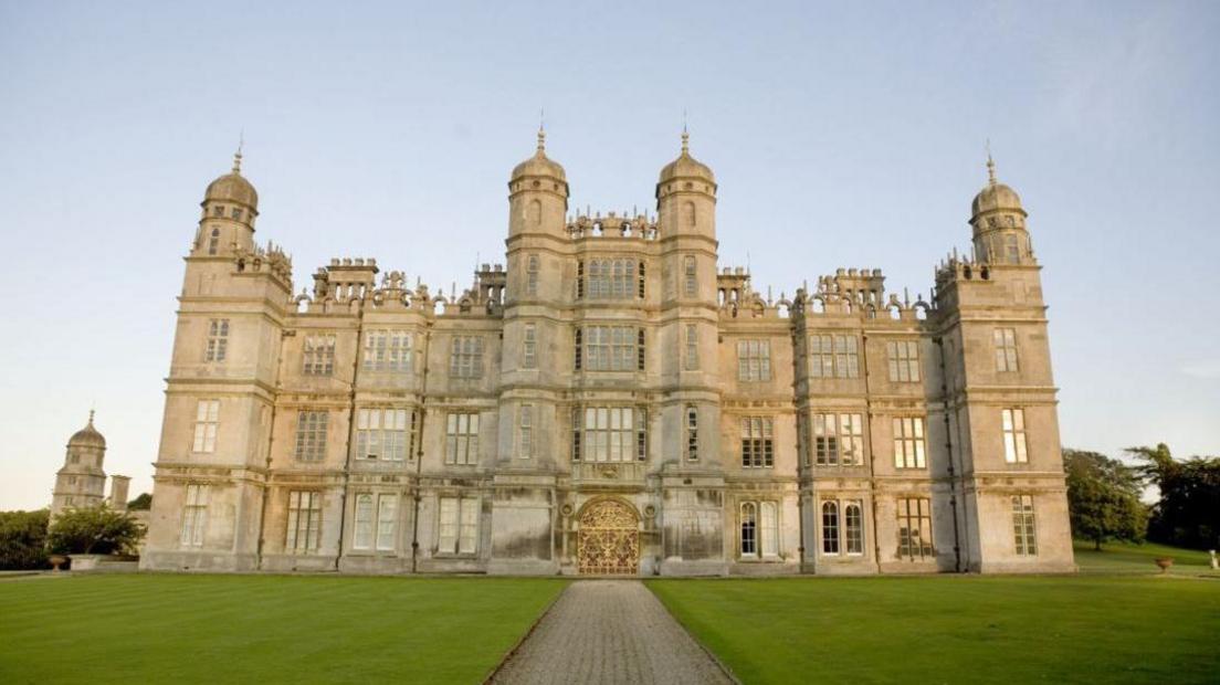 The front of Burghley House, with a pathway leading up to a golden entrance and a neat lawn either side. The building is very grandeur, with several windows on three floors as well as turrets.