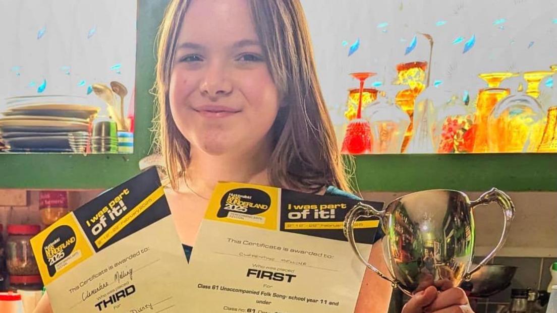 A 13-year-old girl holds two certificates and a trophy and smiles at the camera