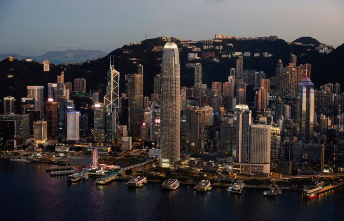 An aerial view of Hong Kong's skyline taken over the harbour. There are boats docked at piers, skyscrapers and lower rise buildings. Mountains can be seen behind and in the distance.