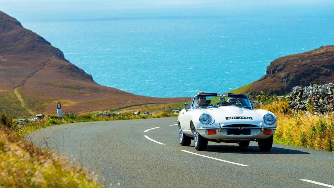 A white classic convertible driving up a hill with coastline cliffs and the turquoise sea in the background.