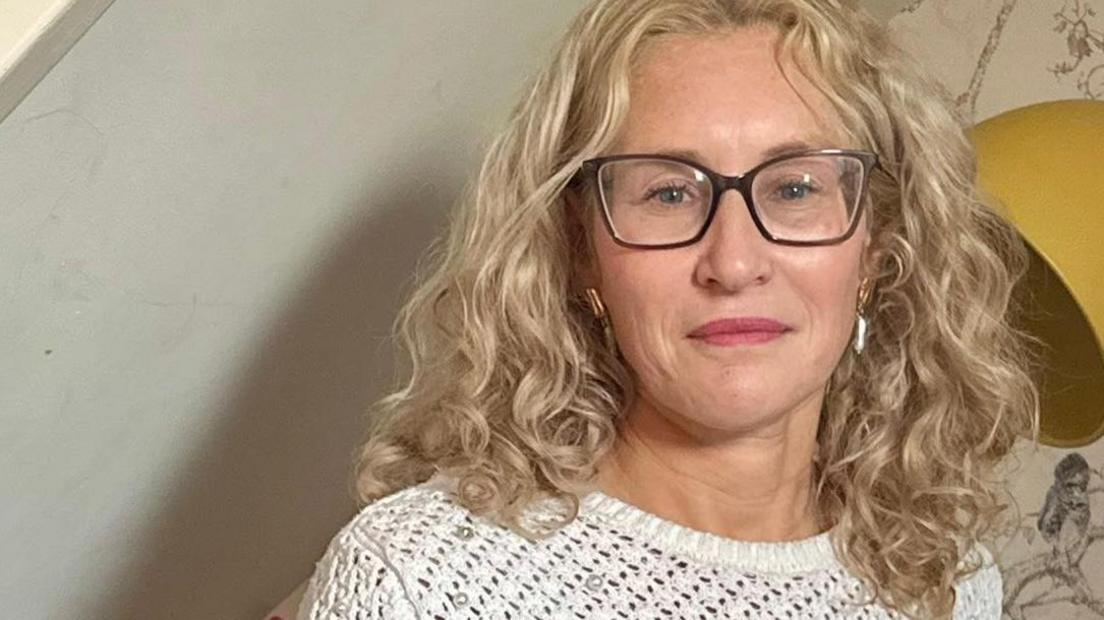 The head and shoulders of a woman with blonde curly hair and glasses, looking at the camera. She wears a white top. A yellow lampshade is behind her, and a book shelf to her left.