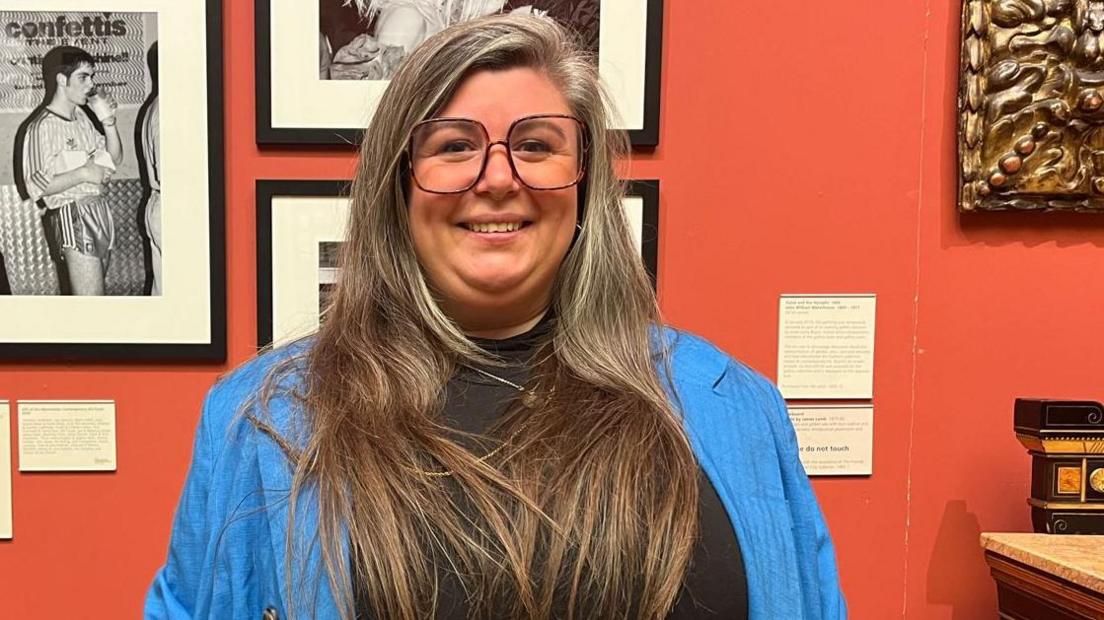Gracie Divall: she has long hair and is wearing large,  black-rimmed glasses, a black top and bright blue shirt. She is standing in the art gallery and smiling at the camera.