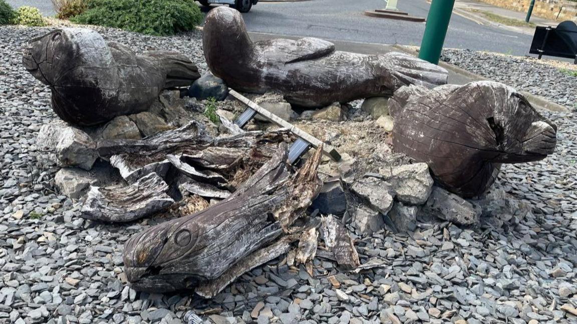 Three weathered brown seal sculptures surround the fragmented rotten remains of the unluckiest seal which has collapsed on itself. They are surrounded by slate chippings on a roundabout.
