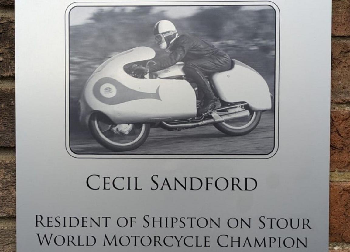 A silver plaque with a picture of a man riding a motorbike. Beneath it says "Cecil Sandford resident of Shipston on Stour, World Motorcycle Champion"