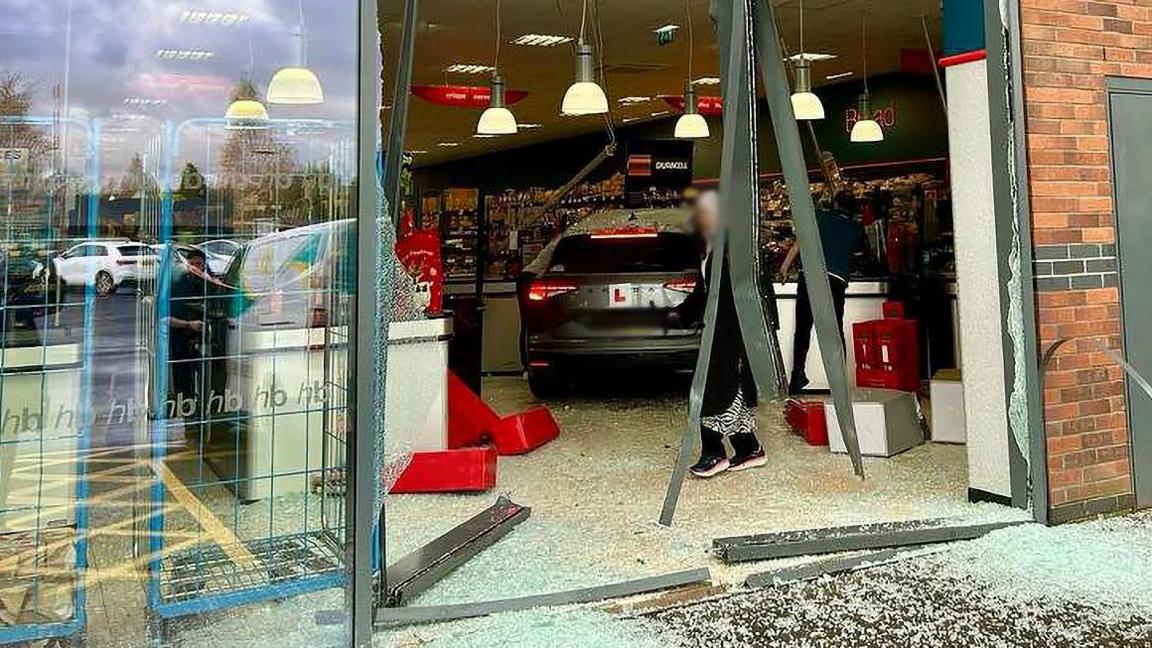 A grey car with an L plate on the back in the middle of carnage, surrounded by smashed glass, tumbled boxes, and hanging metalwork as a woman in a headscarf walks by inside the shop