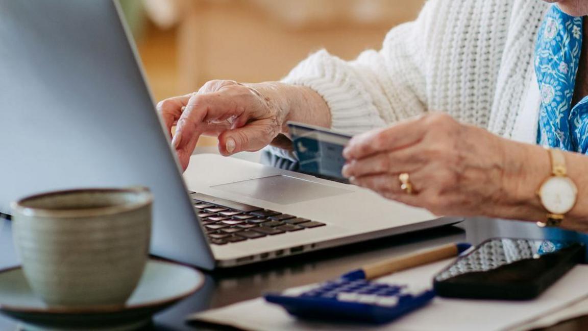 An elderly woman is typing on a laptop with her right hand, and holding a blue bank card in her left. Her face cannot be seen but part of her torso is in view, she is wearing a blue shirt and white cardigan. On the table there is a card reader, a phone and a cup and saucer.