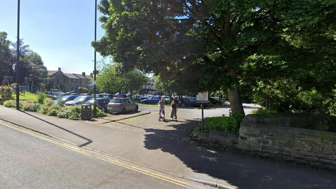 A family walk out of the entrance to a car park in Horsforth Leeds. Several cars are in the car park as well as several trees.