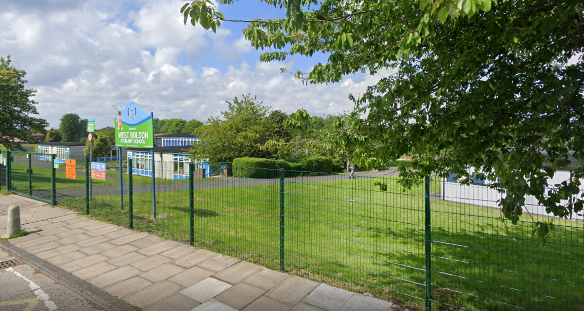 The entrance to the grounds of West Boldon Primary School, which sits behind a wire fence. Bright signage close to green, closed gates gives the name of the school.