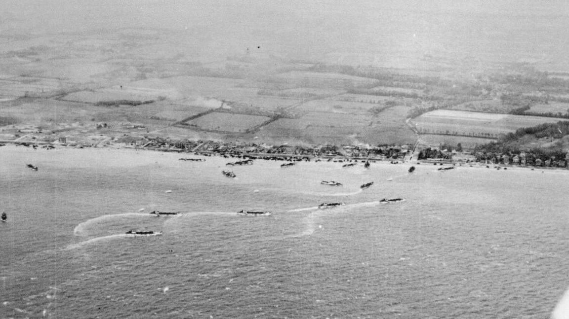 Black and white photo taken from above, looking over sea and coast. In the sea are numerous boats lining up.