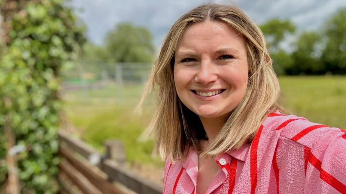 Jessica Featonby smiles as she takes a selfie standing beside a wooden fence in a grassy field. She has shoulder length light hair and wears a pink shirt with red stripes. The background is blurred but you can make out trees and a cloudy sky behind her.