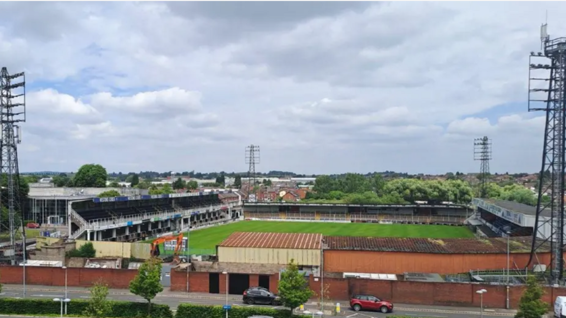 A small football club grounds. Stalls are visible around the pitch and four large pylons can be seen in each corner. There is a road in front of the stadium