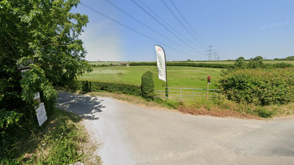 Google Street View image showing the entrance to a farm track from a narrow tarmac lane. A row of large electricity pylons stretch across grassy fields into the distance. A long narrow flag bearing the word "camping" is planted at the entrance to the drive.