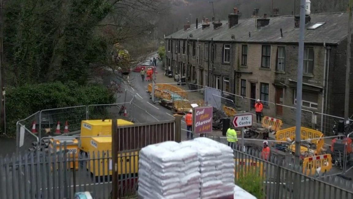 A picture showing a road closed off by road works. Next to the roadworks are a row of houses and on the other side of the road is a business premise.