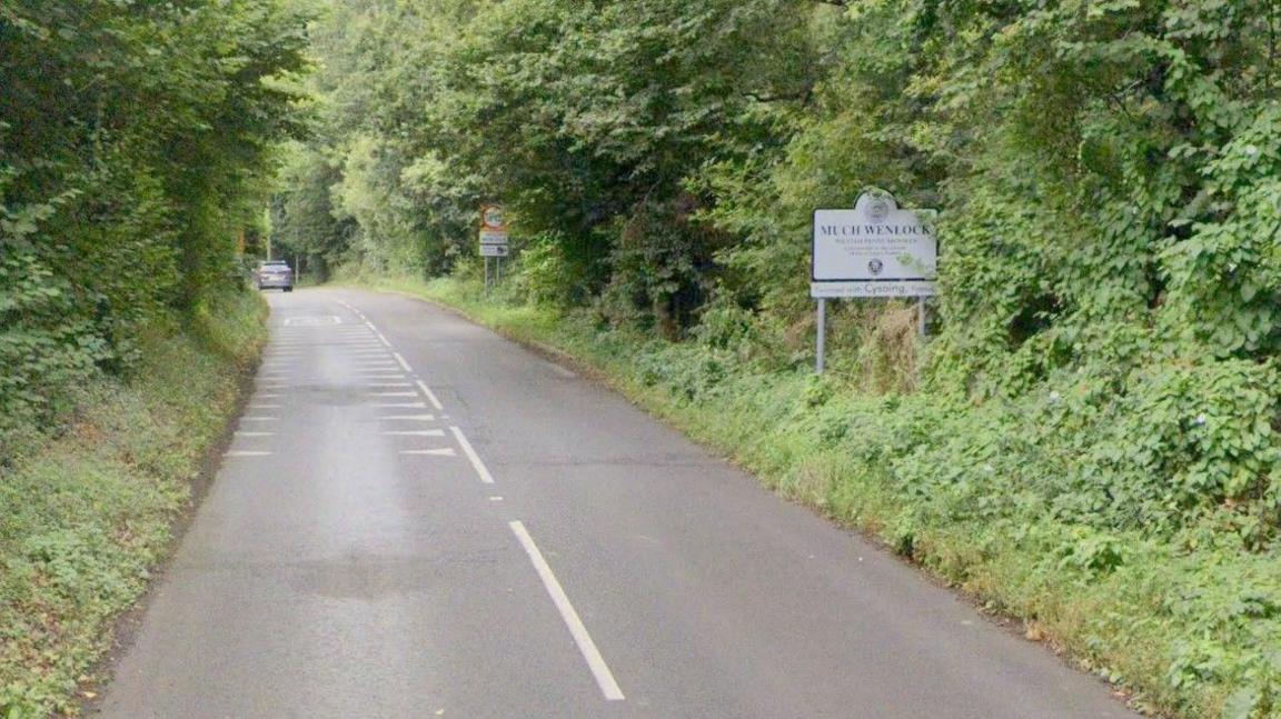 A road surrounded by trees and bushes either side. There is a yellow speed limit sign on the right as well as a white sign which says "Much Wenlock"