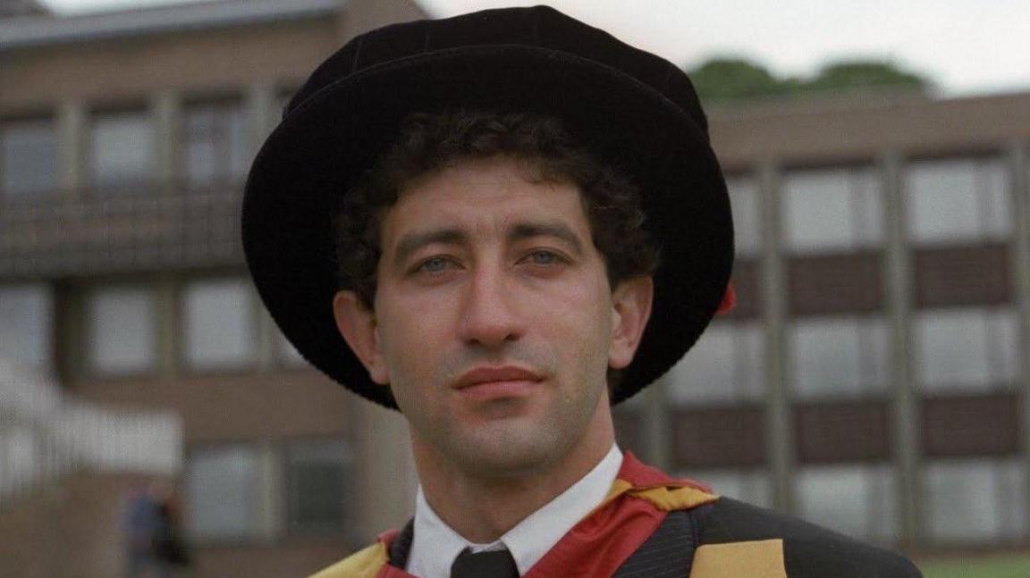 A young man dressed in a graduation gown and hat looking into the camera with a large building behind him. He is wearing a white shirt, black tie and the gown is black, yellow and red.