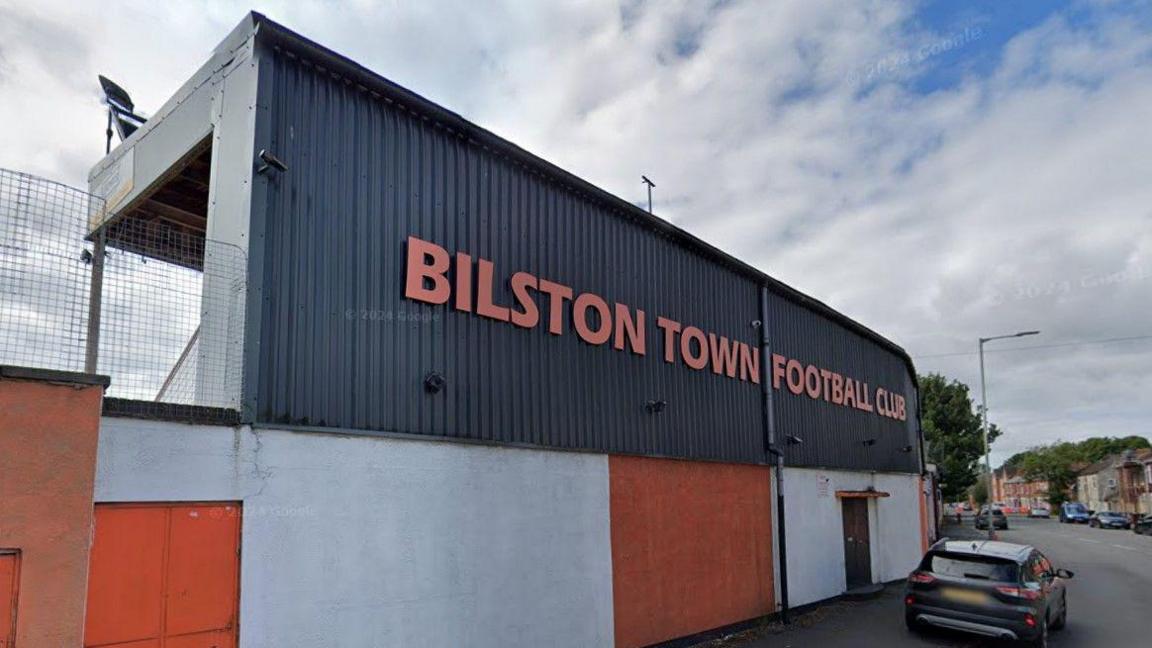 A football stand with red and white panels below a corrugated steel frontage with "Bilston Town Football Club" spelled out in red cut-out lettering. 