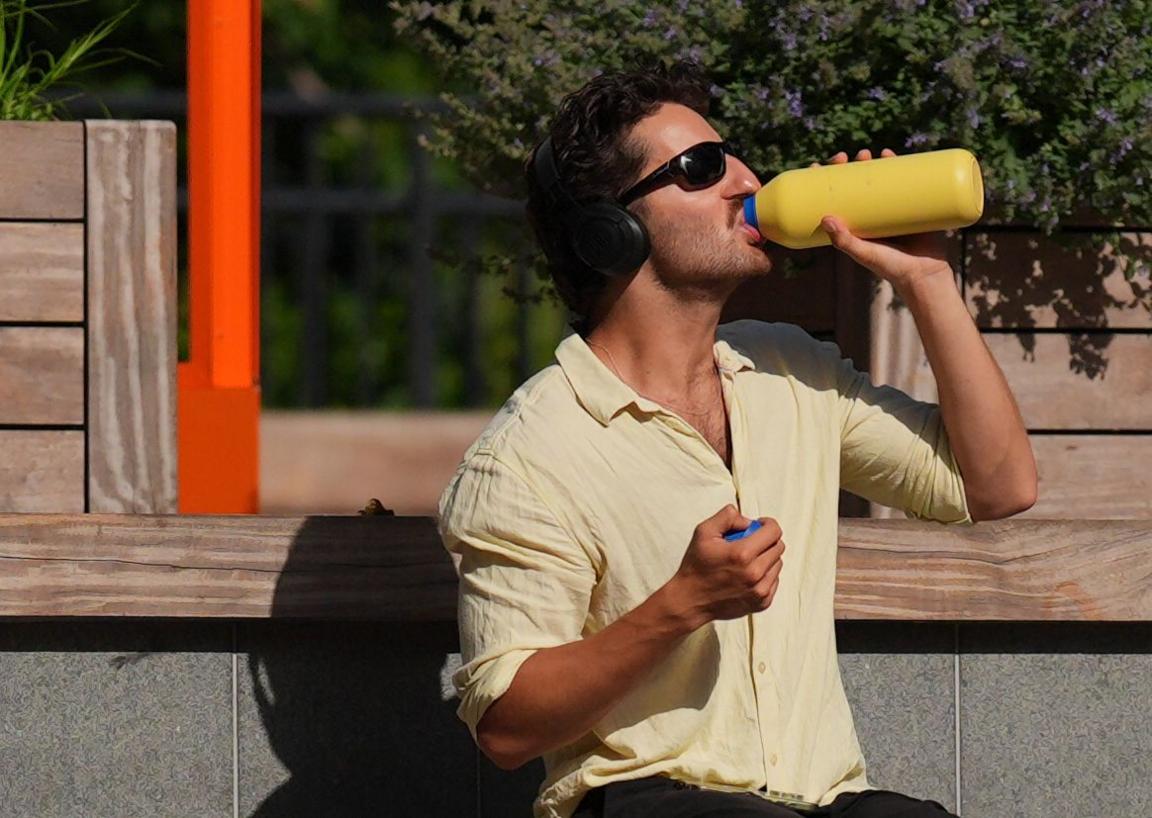 A man in sunglasses and a yellow shirt drinks from a bright yellow water bottle on a bench outside Canary Wharf Station in east London.