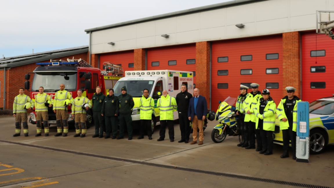 Paramedics, police officers, and firefighters standing in a row outside a fire station and in front of a police car, a fire engine and an ambulance.