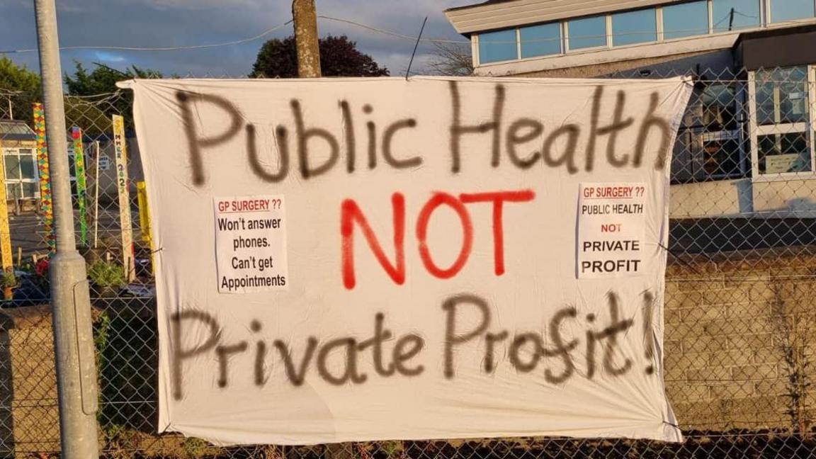 Sheet hung on a fence with the words "Public Health not Private Profit!". Also on the sheet are posters that read "