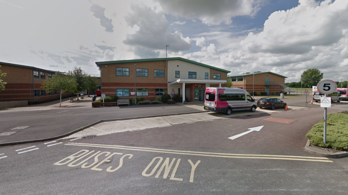 A school building in front of a car park. Words written on the road say buses only, and an arrow points  for the direction of traffic.