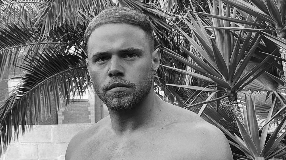 Corey pictured in black and white. He is bare chested and standing in front of a wall and a palm tree.