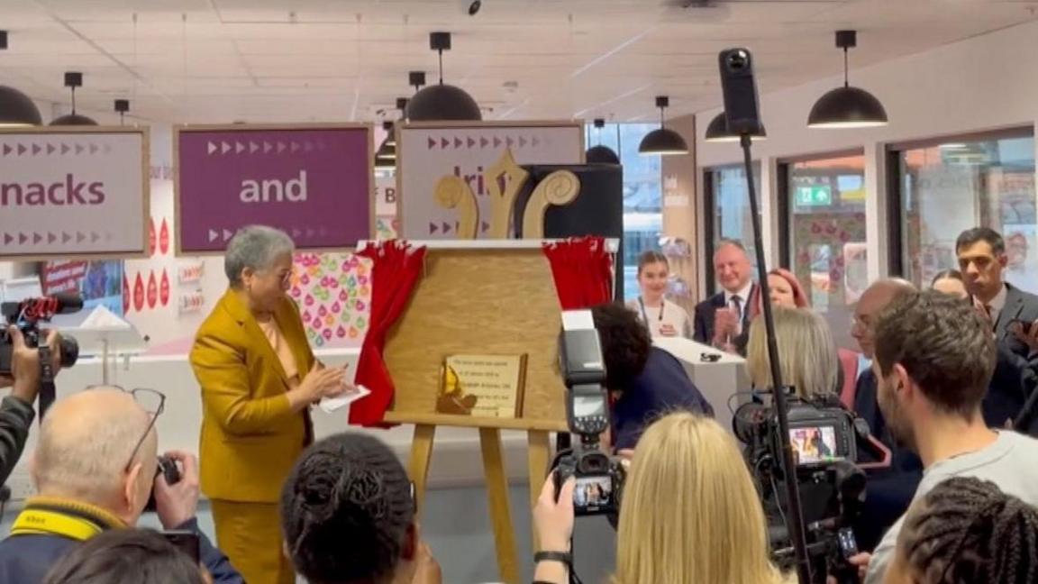 A screengrab from a video of Professor Dame Elizabeth Anionwu wearing a mustard yellow suit and stood, mid-clap, looking at her plaque, which has just been unveiled behind red curtains. People are stood around her with cameras and also clapping. 