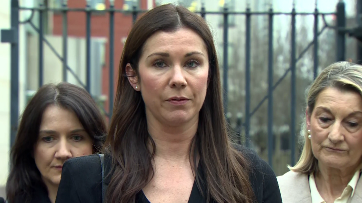 A group stand outside court. One woman is standing in front and is speaking to the media. She has long brown hair and is dressed in black. 