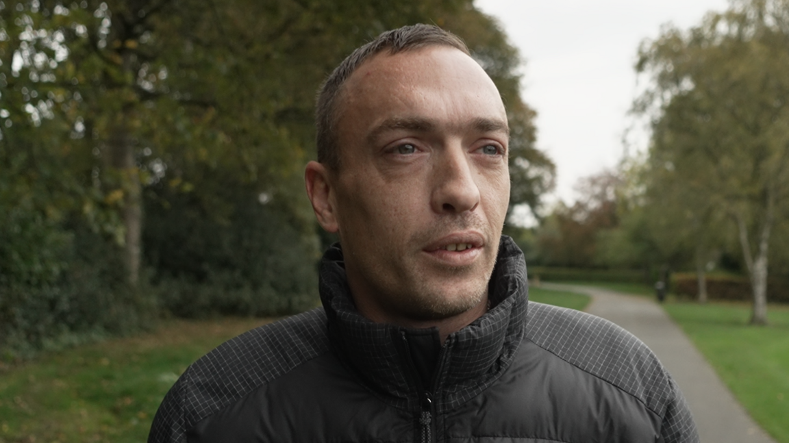 A man in a black coat stands on a path in a park, surrounded by green grass and trees. He has an inquisitive expression, looking slightly beyond the camera.
