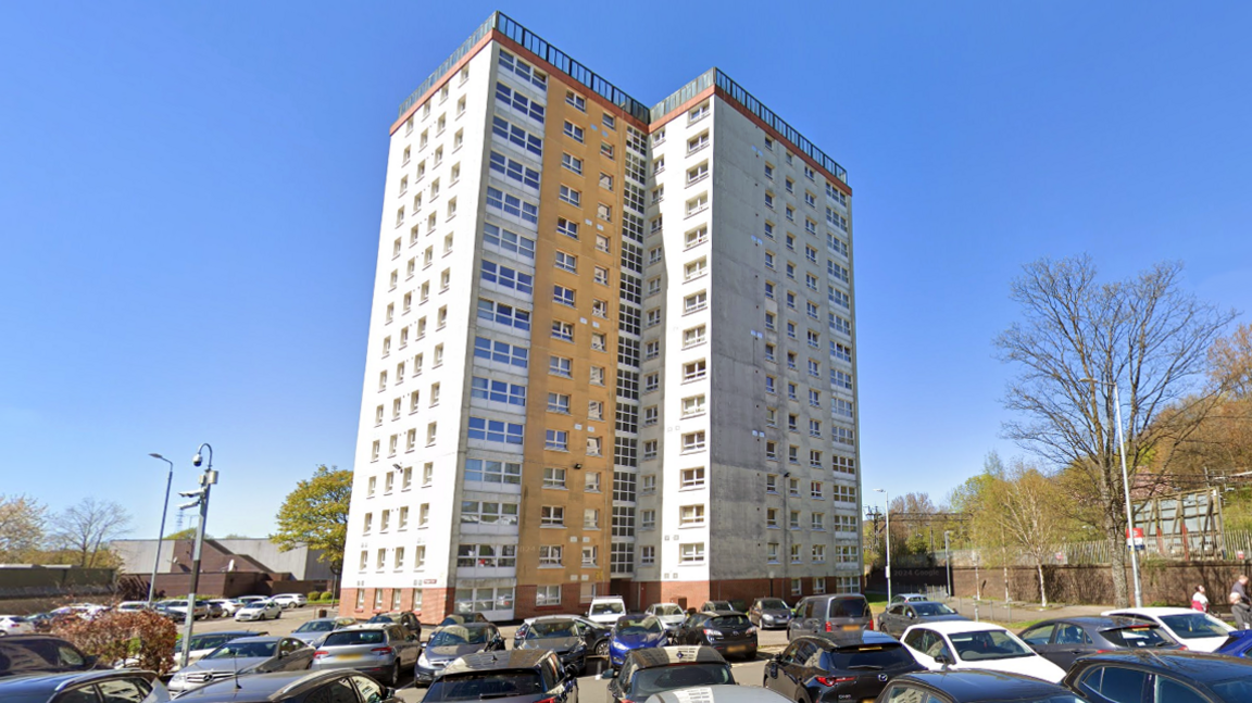 A 15 floor tower block rises high into the blue sky with numerous cars parked at the bottom.