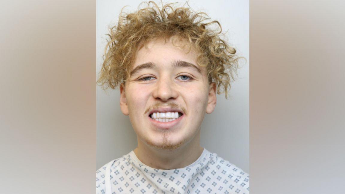 Teddy Kelt smiles in his police custody photo. He has blonde curly hair. He is wearing a hospital gown. 
