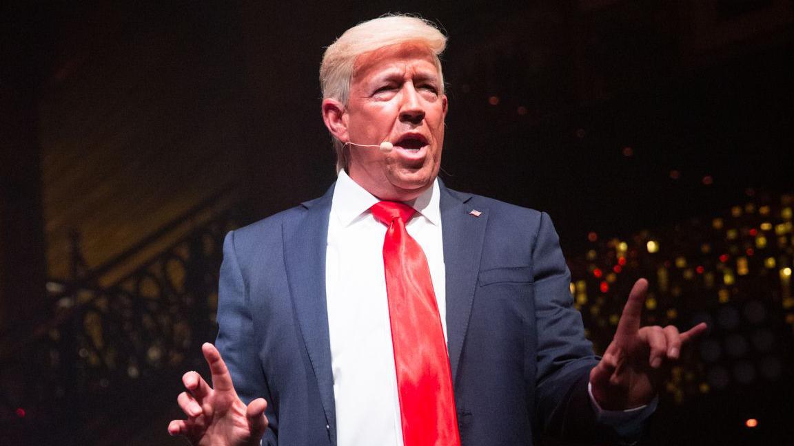 Mike Osman dressed as Donald Trump. He is wearing a blonde wig, a blue suit and red tie.