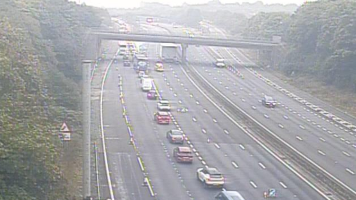 A queue of cars and lorries on four lanes of a southbound motorway with a bridge crossing the road and clear crarriageways on the other side of the central reservation.