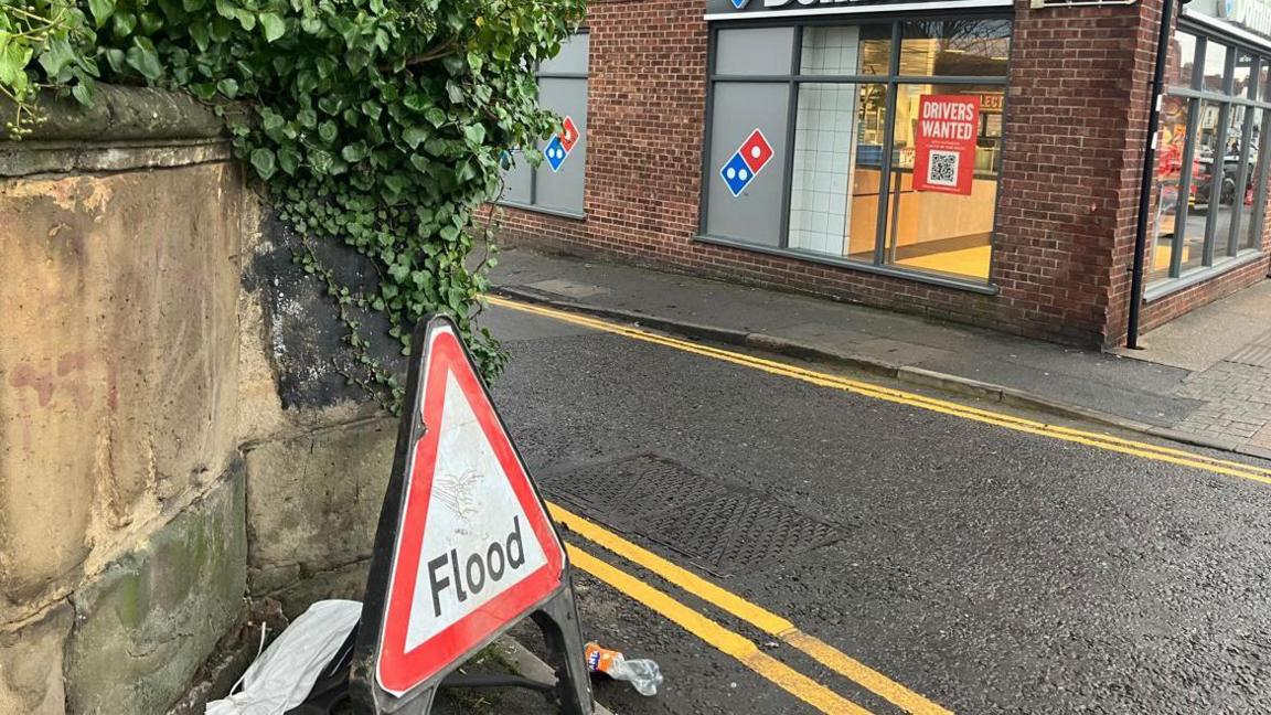 A triangular red and white flood sign at the end of the street. There are double yellow lines on both sides of the street and there is a Domino's on the opposite corner