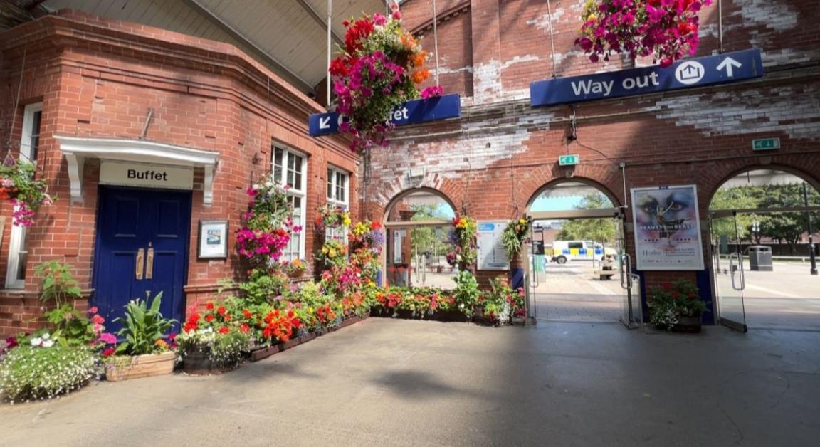 Several colourful plants dotted around the red-brick walls and arched entranceways of Bridlington station with some hanging plants near a "way out" sign