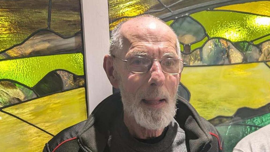 Melvin looking into the camera, stood in front of a large stained glass window. Melvin wears glasses and a black jacket over a grey jumper.